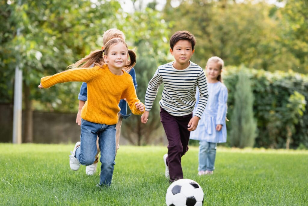 Cheerful multiethnic kids playing football near friends on blurr
